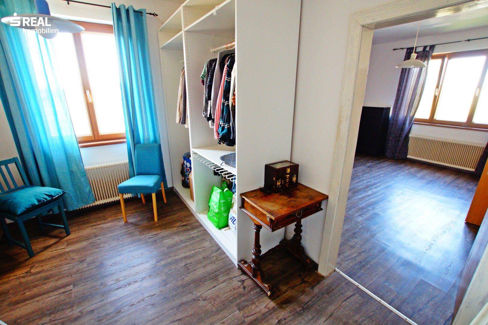 Schlafzimmer mit offenem Kleiderschrank, blauen Stühlen und Zugang zu einem weiteren Raum.
