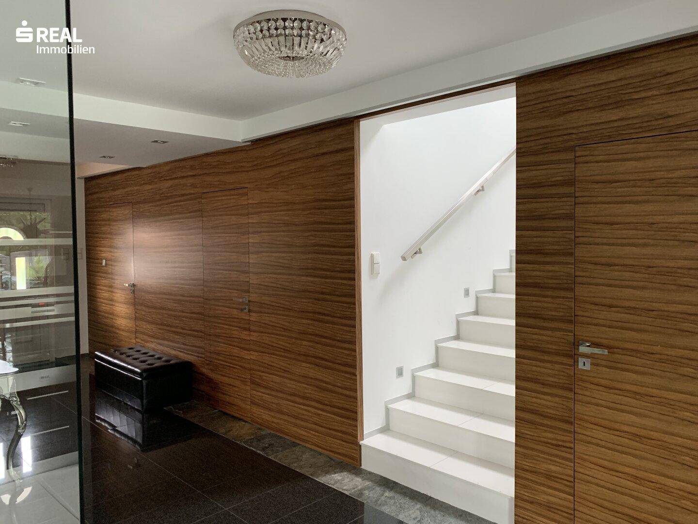 Heller Flur mit Holzvertäfelung, weißer Treppe und modernem Kronleuchter.