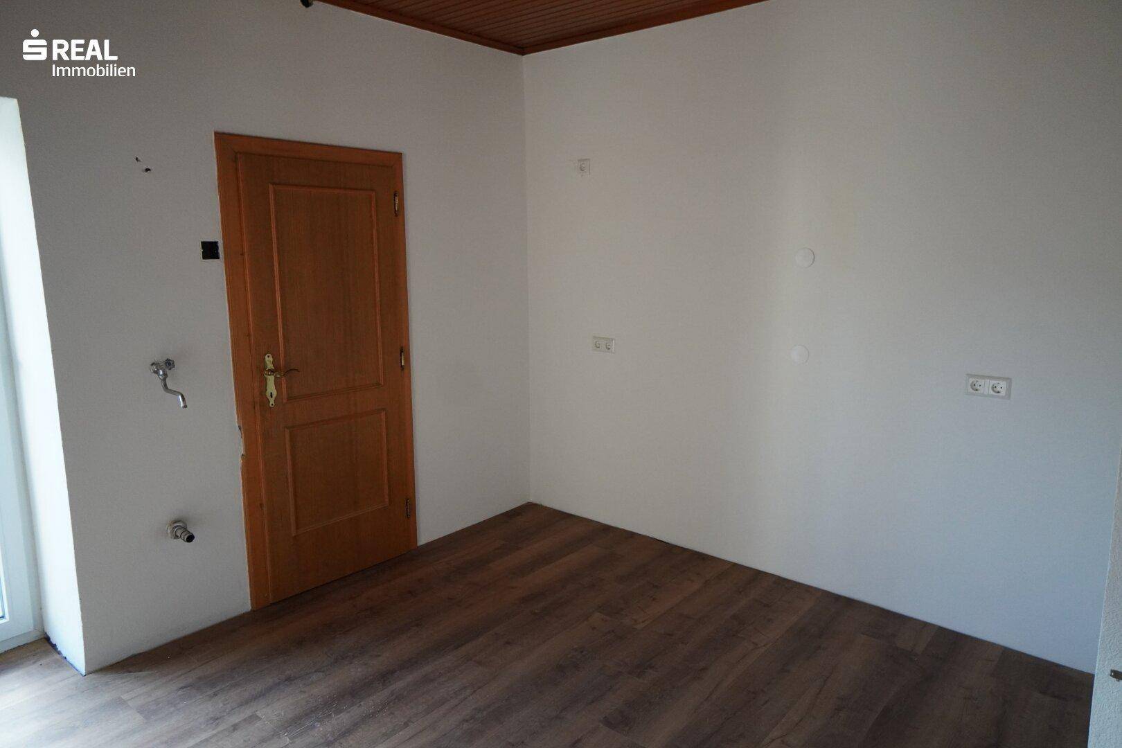 Leerer Küchenraum mit Holzdecke und Anschlüssen für eine zukünftige Einbauküche.
