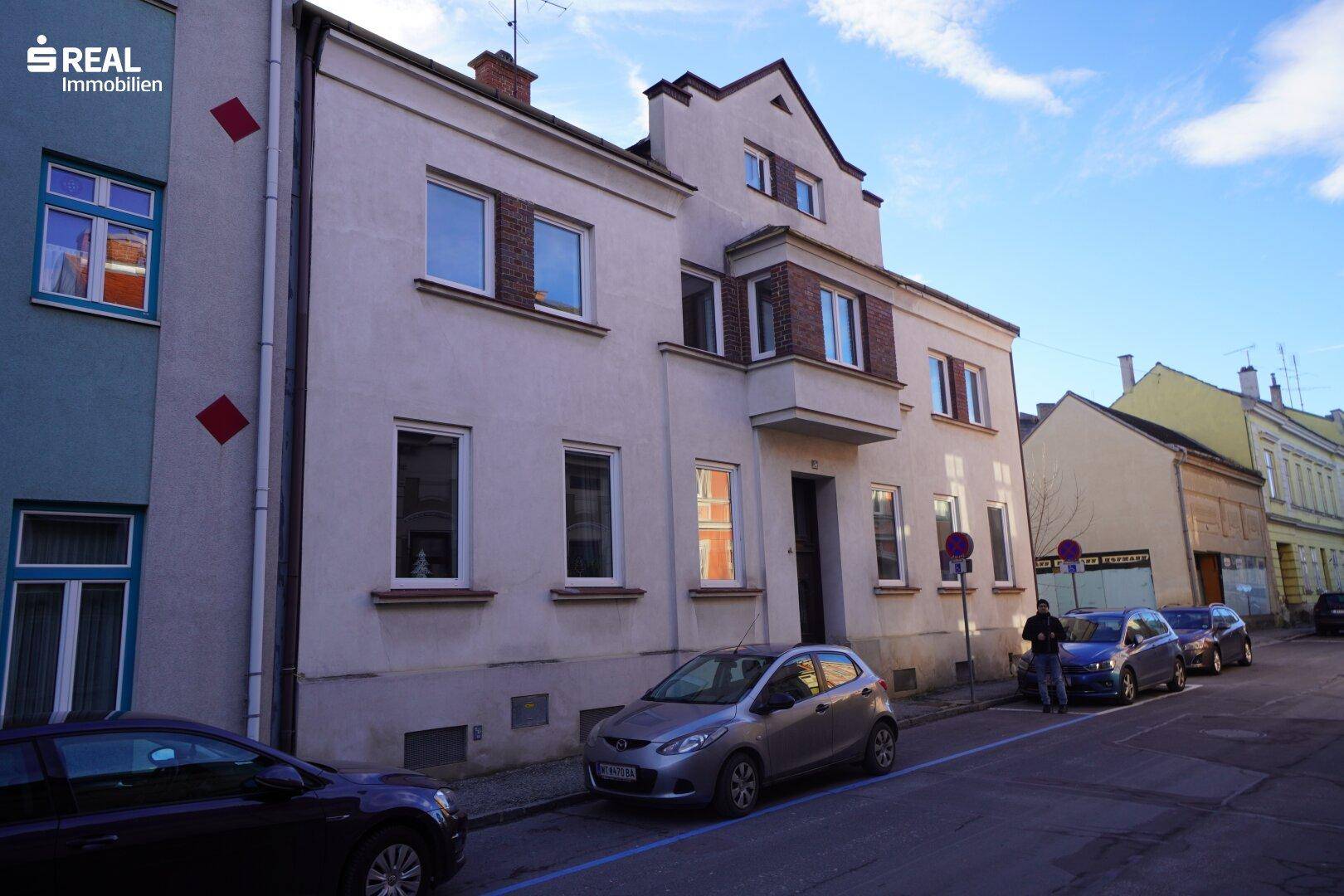 Fassade eines Altbaus an einer belebten Straße mit parkenden Fahrzeugen.
