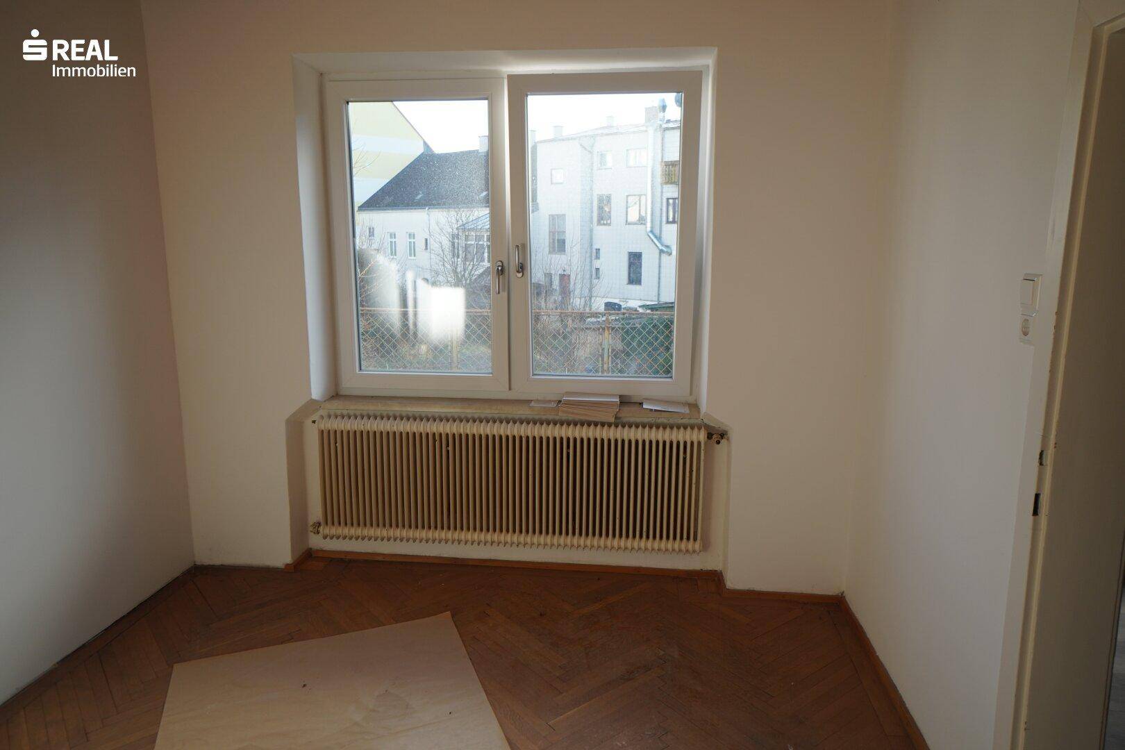 Helles Schlafzimmer mit großem Fenster, Heizkörper und traditionellem Parkettboden.