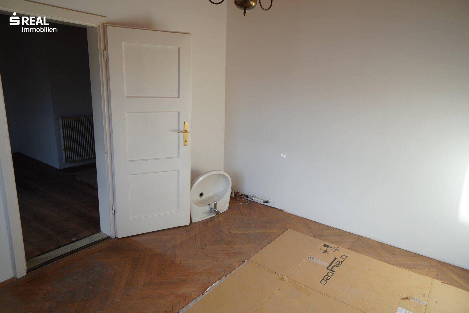 Leeres Schlafzimmer mit Fischgrätparkett und einer Tür, die zu einem weiteren Raum führt.