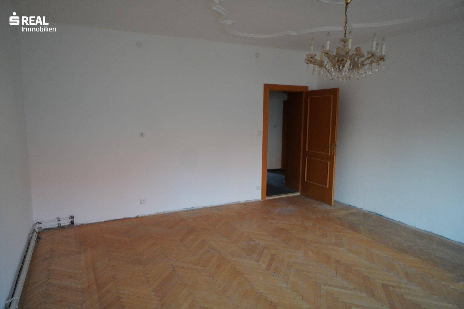 Geräumiges Wohnzimmer mit Parkettboden, Kronleuchter und einer verzierten Stuckdecke.