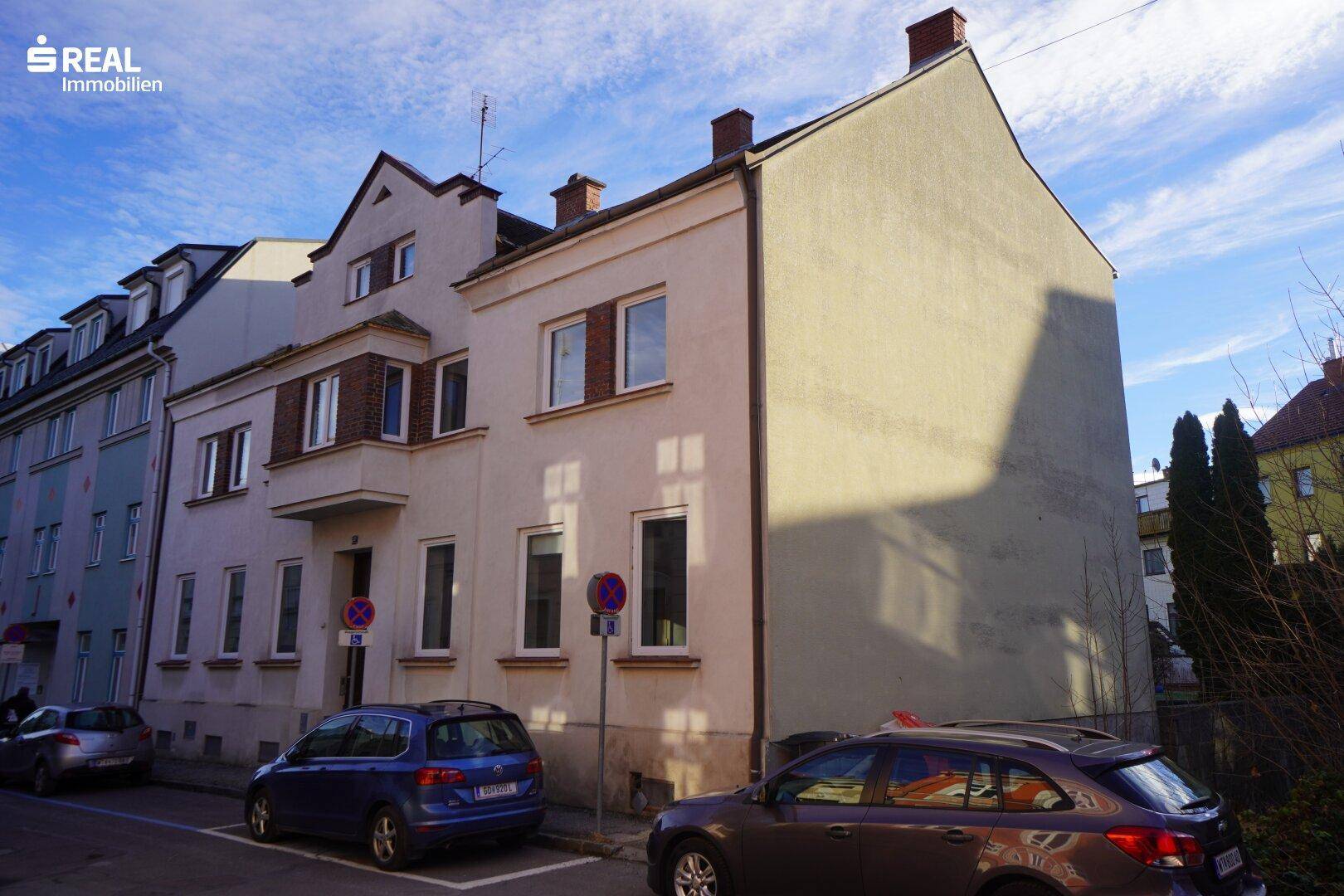 Historisches Gebäude an einer Straße mit parkenden Autos und blauer Himmelskulisse.