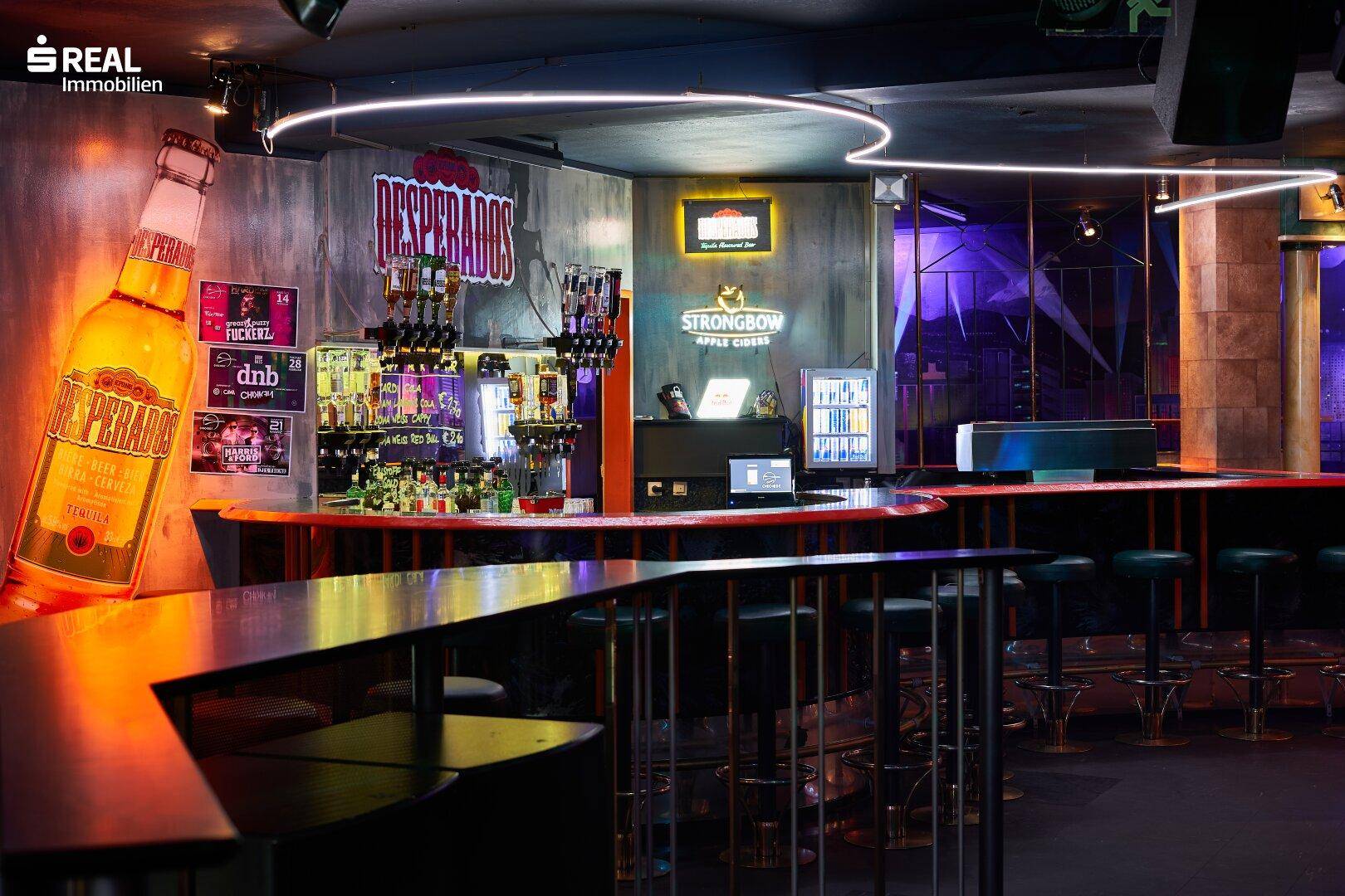 Detailansicht einer Bar mit Flaschenregalen, Neonlicht und Werbeschildern.