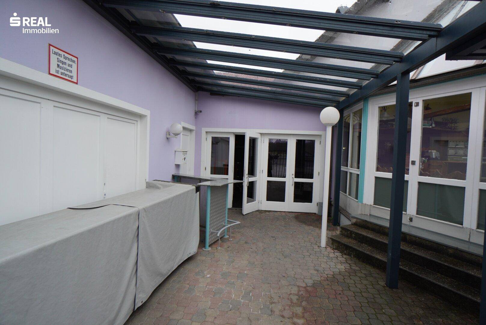 Überdachter Eingangsbereich mit gepflastertem Boden und Zugangstüren zum Gebäude.