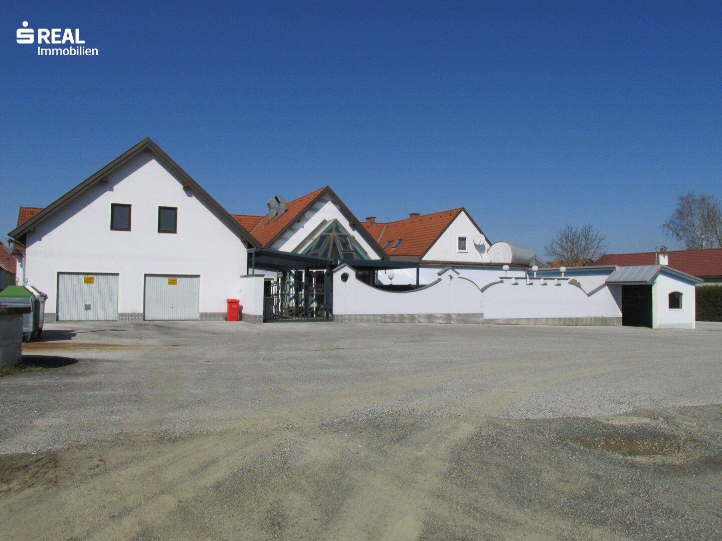 Gebäudefassade mit Garagen, Glasdach und großem Vorplatz unter blauem Himmel.
