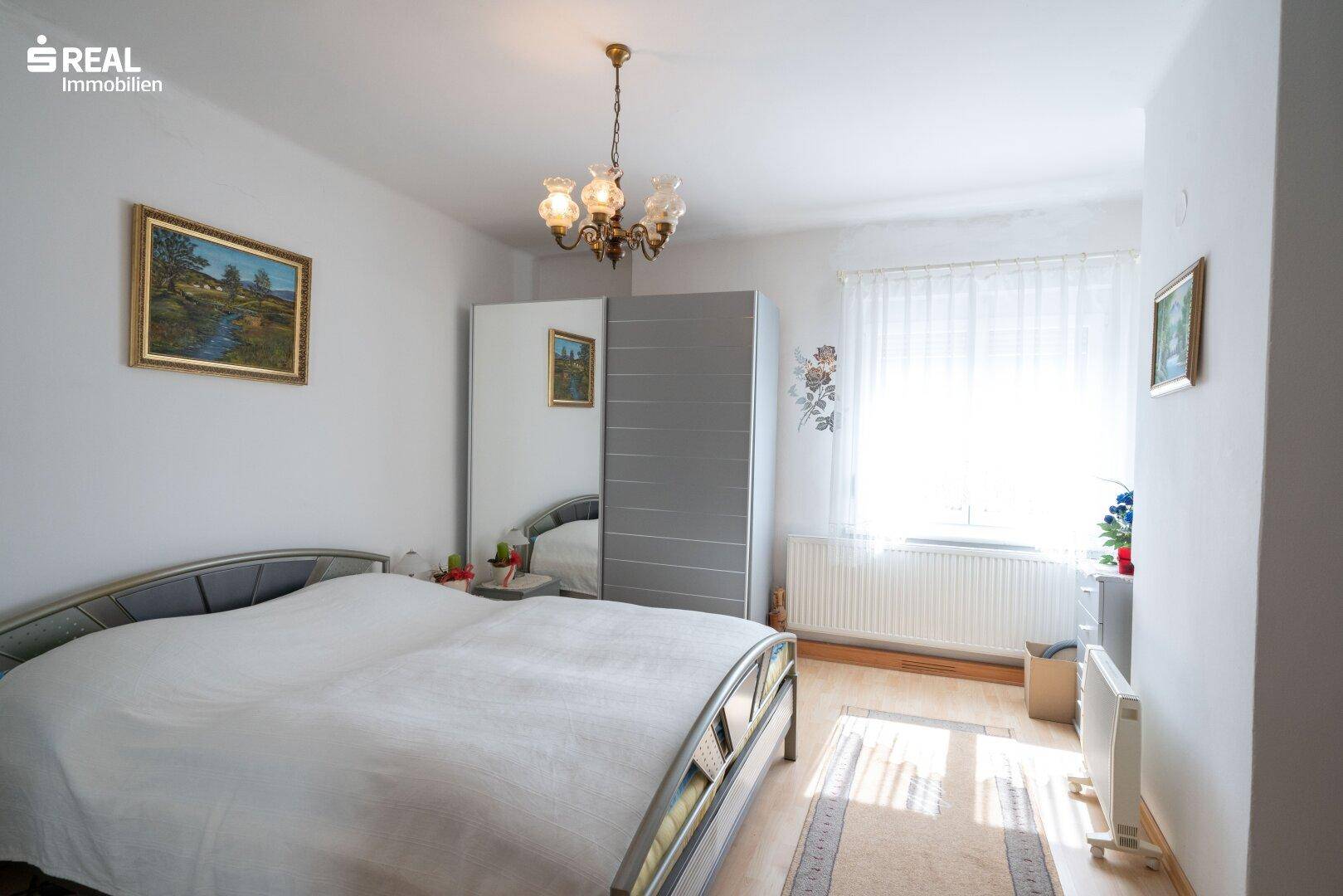 Helles Schlafzimmer mit Doppelbett, Kleiderschrank und großem Fenster.