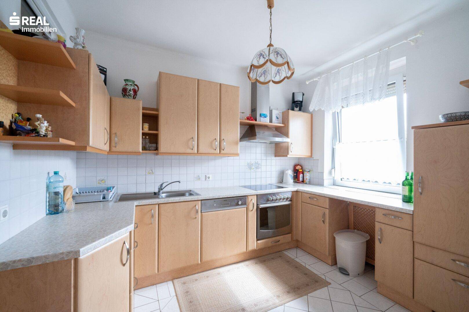 Funktionale Küche mit hellen Holzschränken, Fliesenboden und einem Fenster über der Spüle.