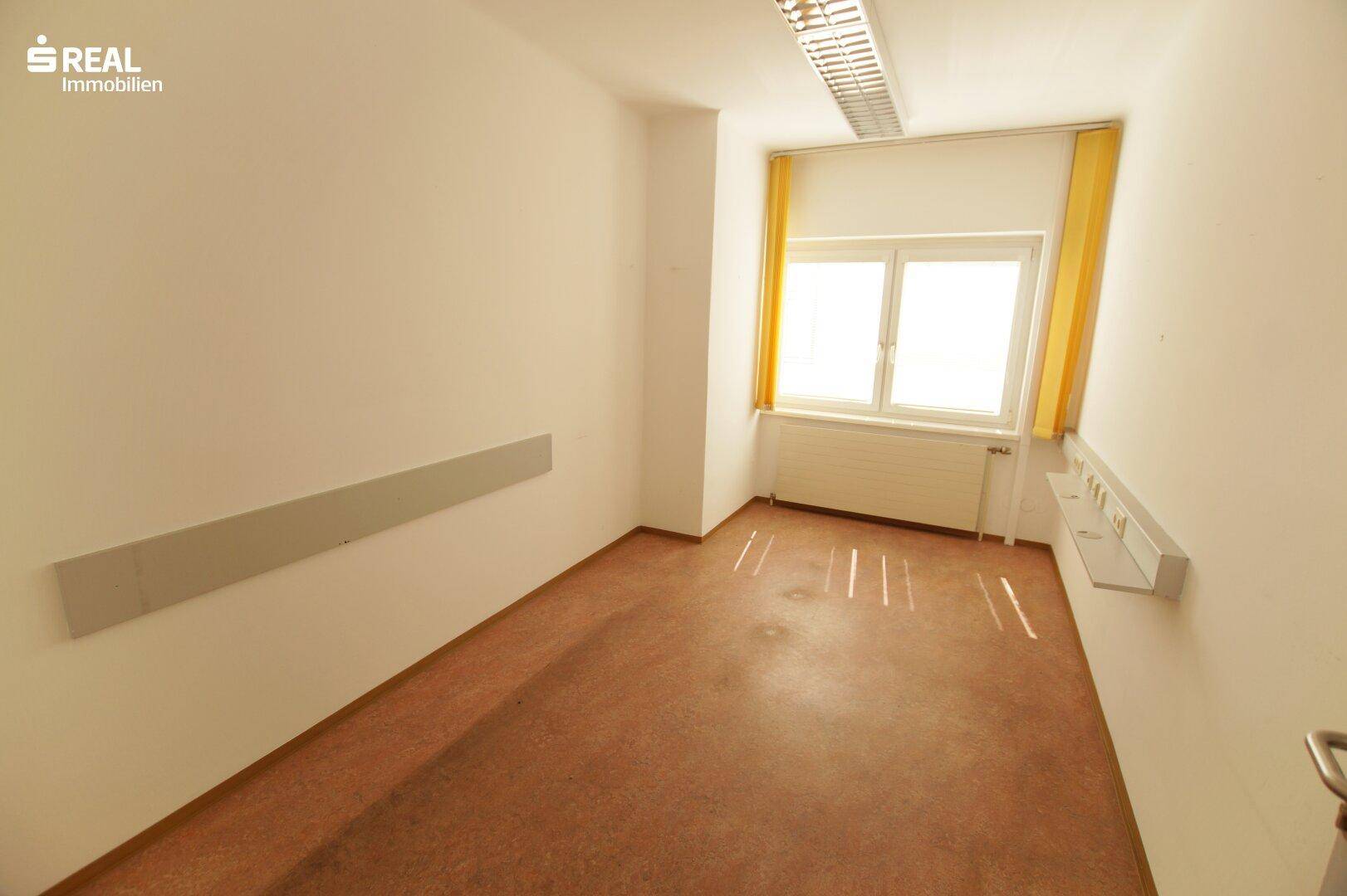 Heller Büroraum mit Fenster, gelben Vorhängen und braunem Bodenbelag.