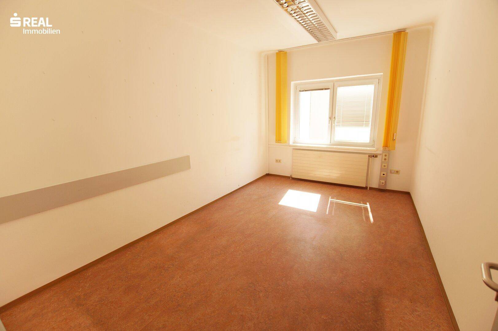 Heller Büroraum mit Fenster, gelben Vorhängen und braunem Bodenbelag.
