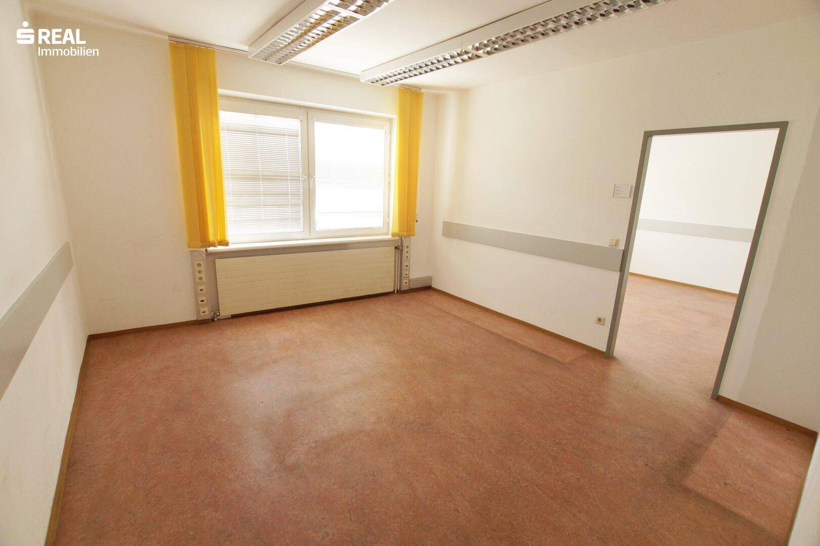 Heller Büroraum mit Fenster, gelben Vorhängen und braunem Bodenbelag.
