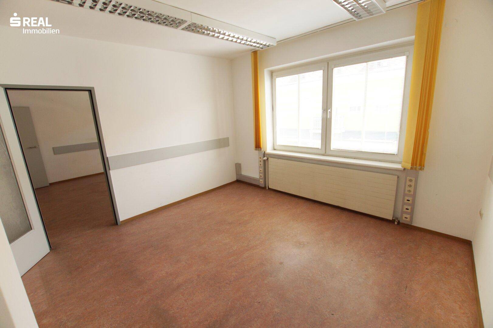 Heller Büroraum mit Fenster, Deckenbeleuchtung und braunem Bodenbelag.