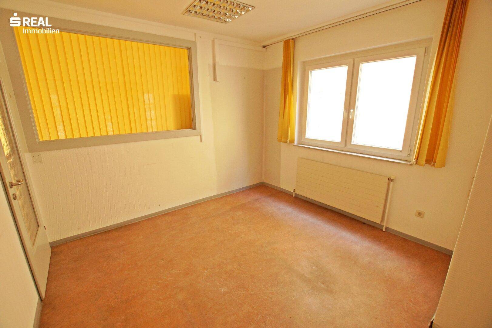 Leerer Raum mit hellem Boden, Fenster mit gelben Vorhängen und Heizkörper unter dem Fenster.