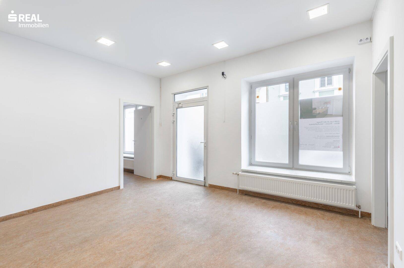 Geräumiger Büroraum mit großen Fenstern, viel Tageslicht und einem Heizkörper unter dem Fenster.
