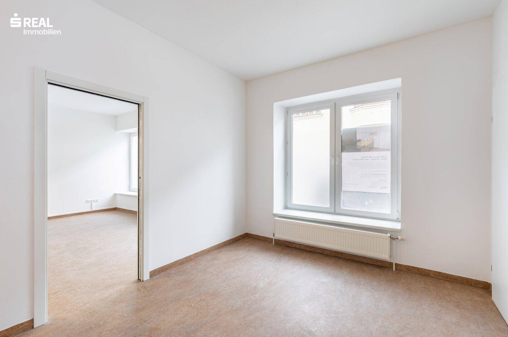 Heller Raum mit großem Fenster und Heizkörper, bietet Zugang zu einem angrenzenden Bereich.