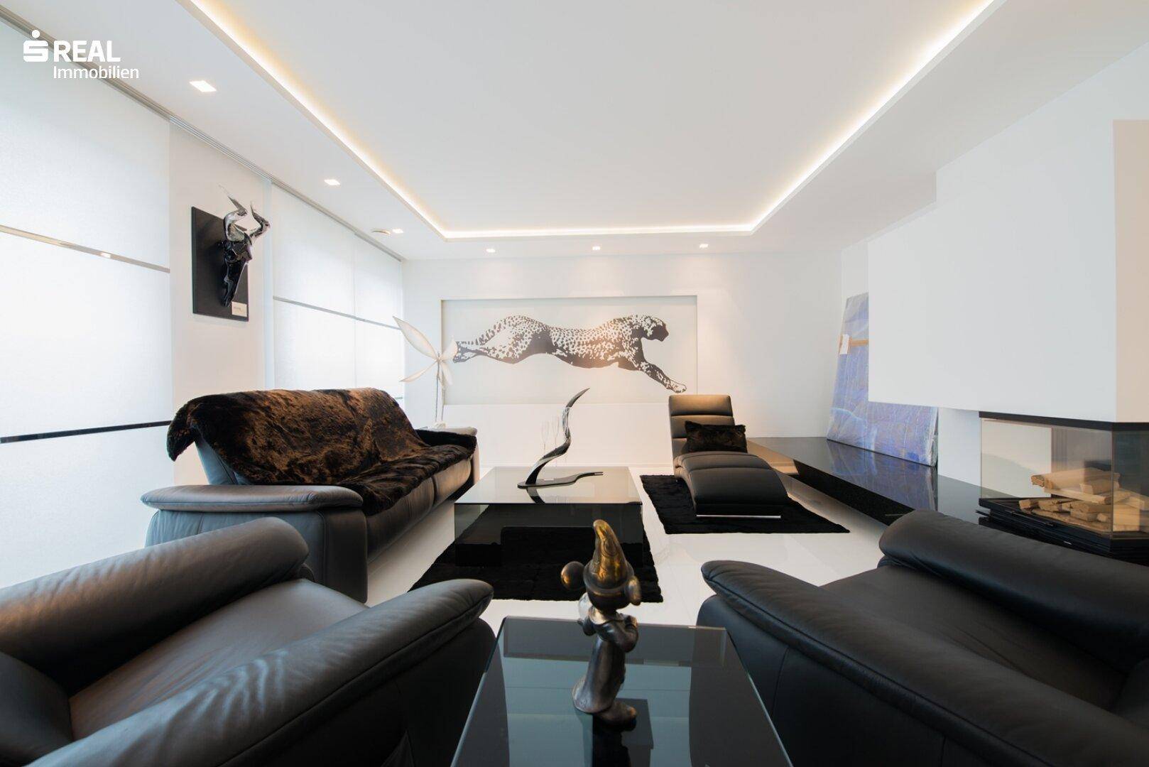 Elegantes Wohnzimmer mit schwarzen Ledersofas, modernem Kamin und indirekter Deckenbeleuchtung.