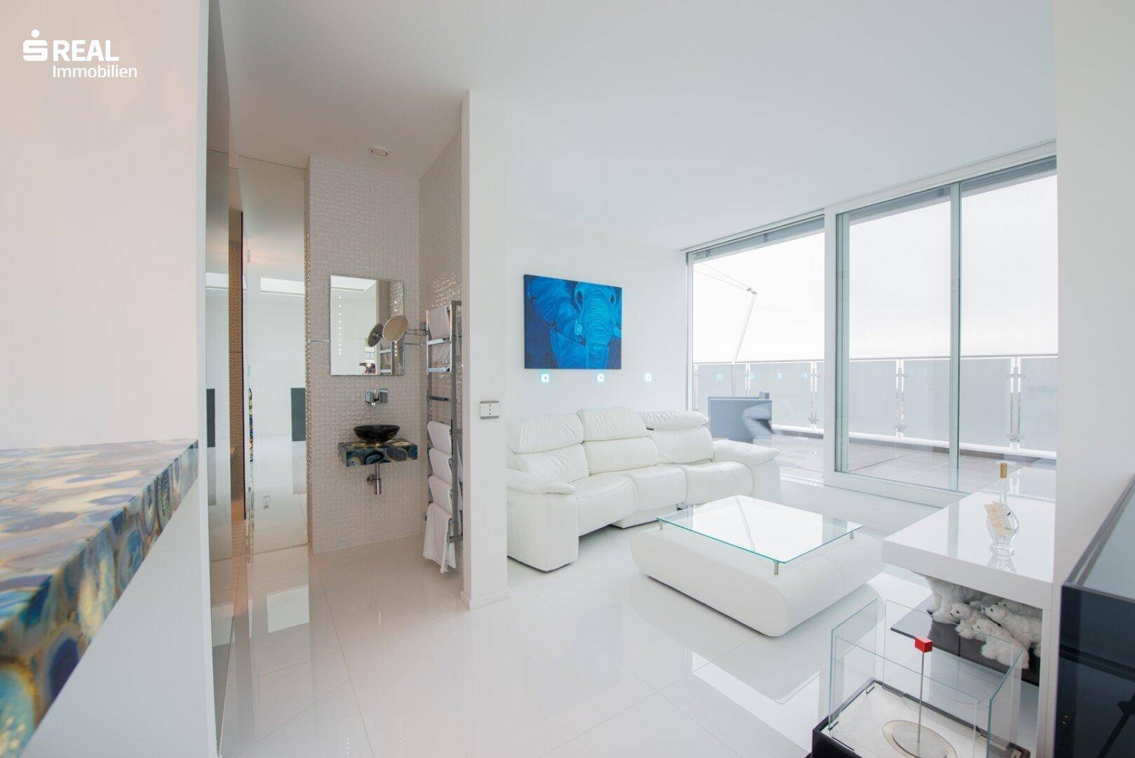 Helles Wohnzimmer mit weißem Sofa, großen Fenstern und Zugang zu einer Außenterrasse.
