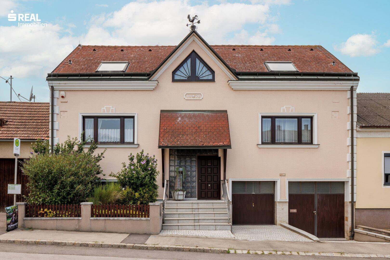 Traditionelles Wohnhaus mit Satteldach, Gaubenfenstern und zwei Garagen im Erdgeschoss.