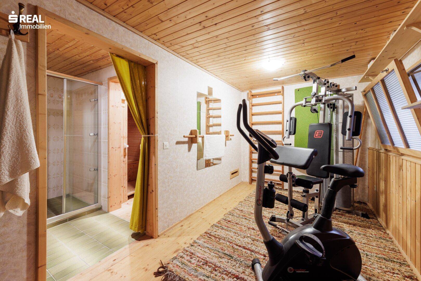 Fitnessraum mit Trainingsgeräten, Holzboden und Zugang zu einer Duschkabine.