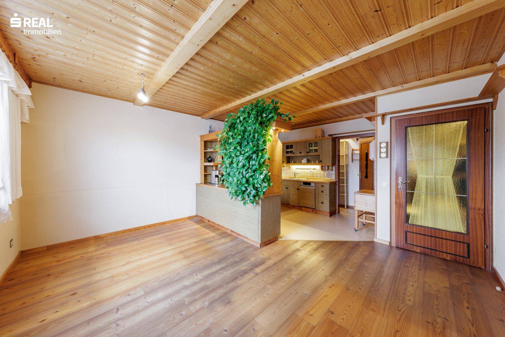 Geräumiges Wohnzimmer mit Holzdielenboden, Holzbalkendecke und offenem Übergang zur Küche.