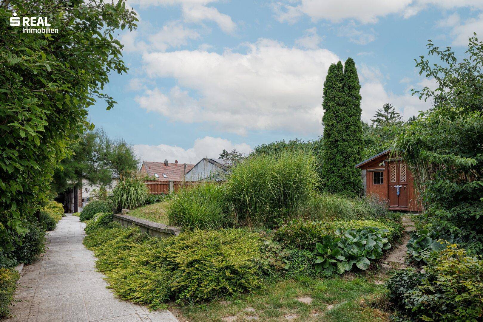 Geschwungener Gartenweg umgeben von üppiger Vegetation und einem kleinen Gartenhaus im Hintergrund.