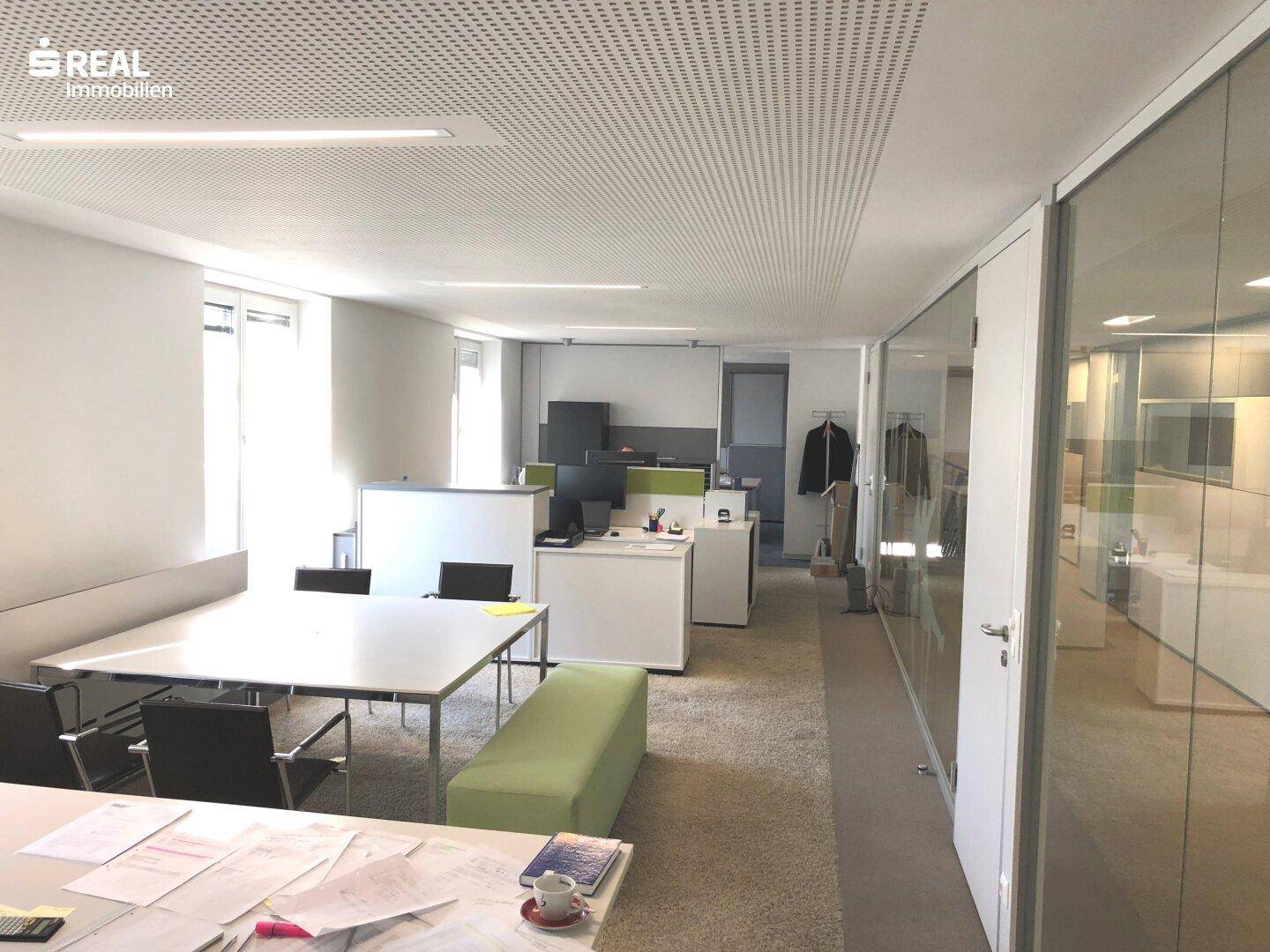 Großes, offenes Büro mit mehreren Arbeitsplätzen, Teppichboden und viel natürlichem Licht durch Fenster.