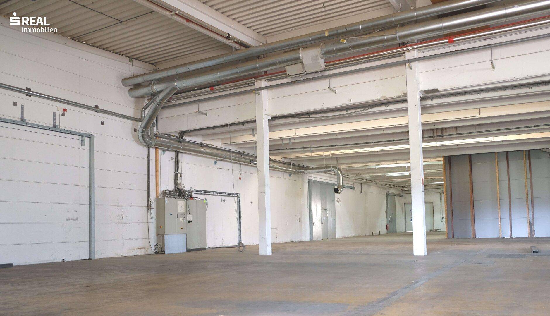 Geräumige Lagerhalle mit hohen Decken, Betonboden und sichtbaren Lüftungsrohren.