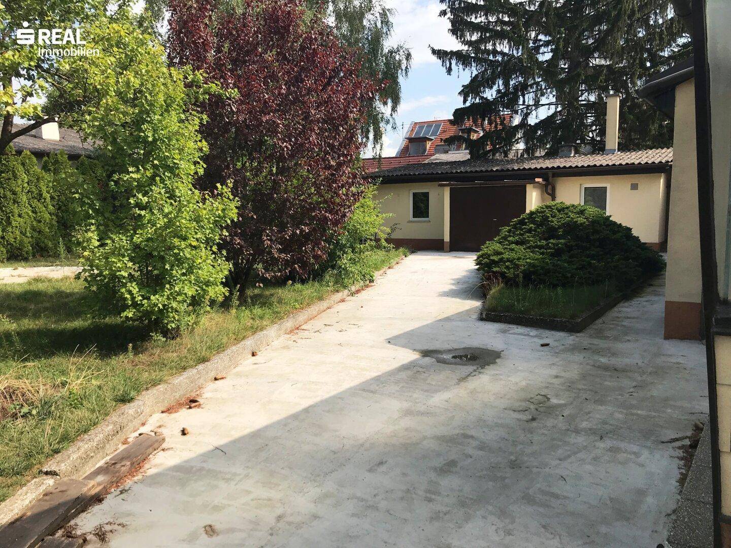 Betonierte Auffahrt führt zur Garage, umgeben von Rasenflächen und Bäumen.
