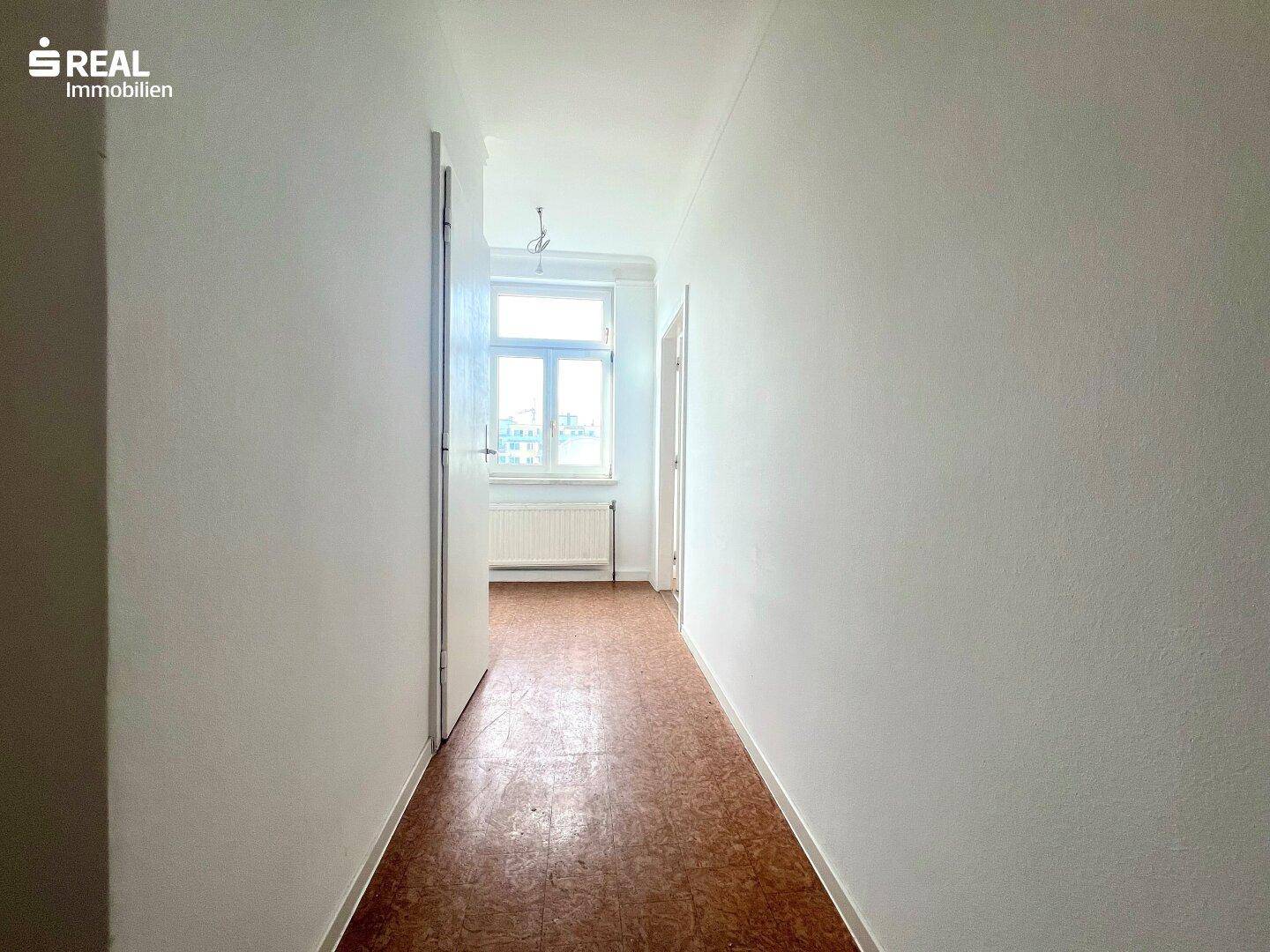 Heller Flur mit Fenster am Ende, braunem Bodenbelag und weißen Wänden.