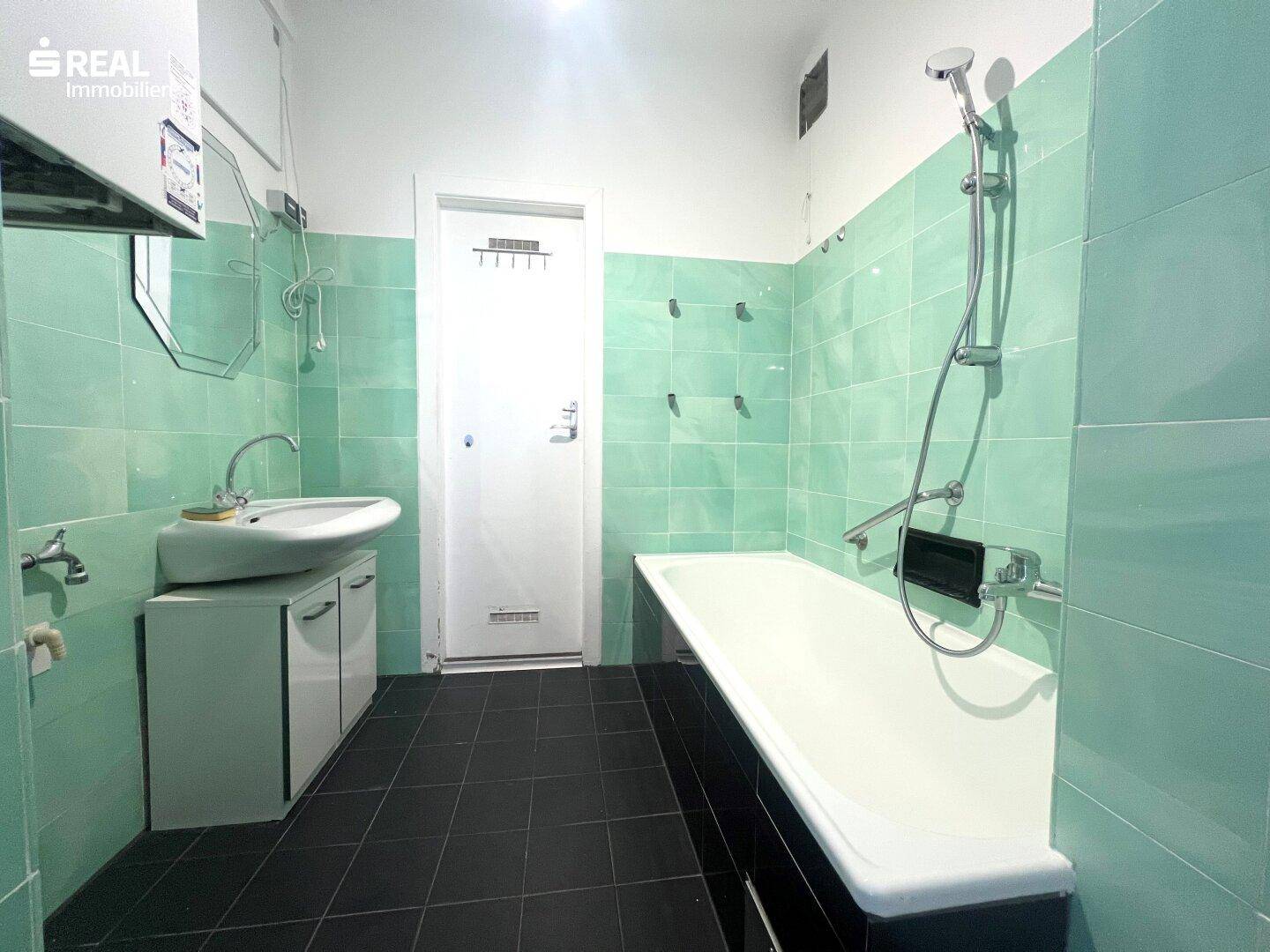 Badezimmer mit grünen Wandfliesen, schwarzem Boden und Badewanne mit Duschfunktion.
