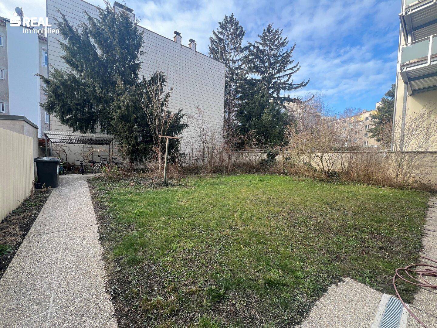 Grüner Gartenbereich mit Rasenfläche, Bäumen und einem gepflasterten Weg hinter dem Gebäude.
