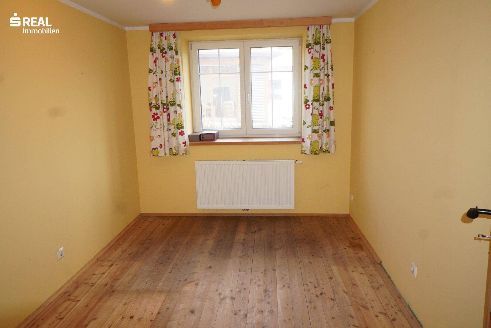 Helles Zimmer mit Holzboden, gelben Wänden und Fenster mit Blick ins Grüne.
