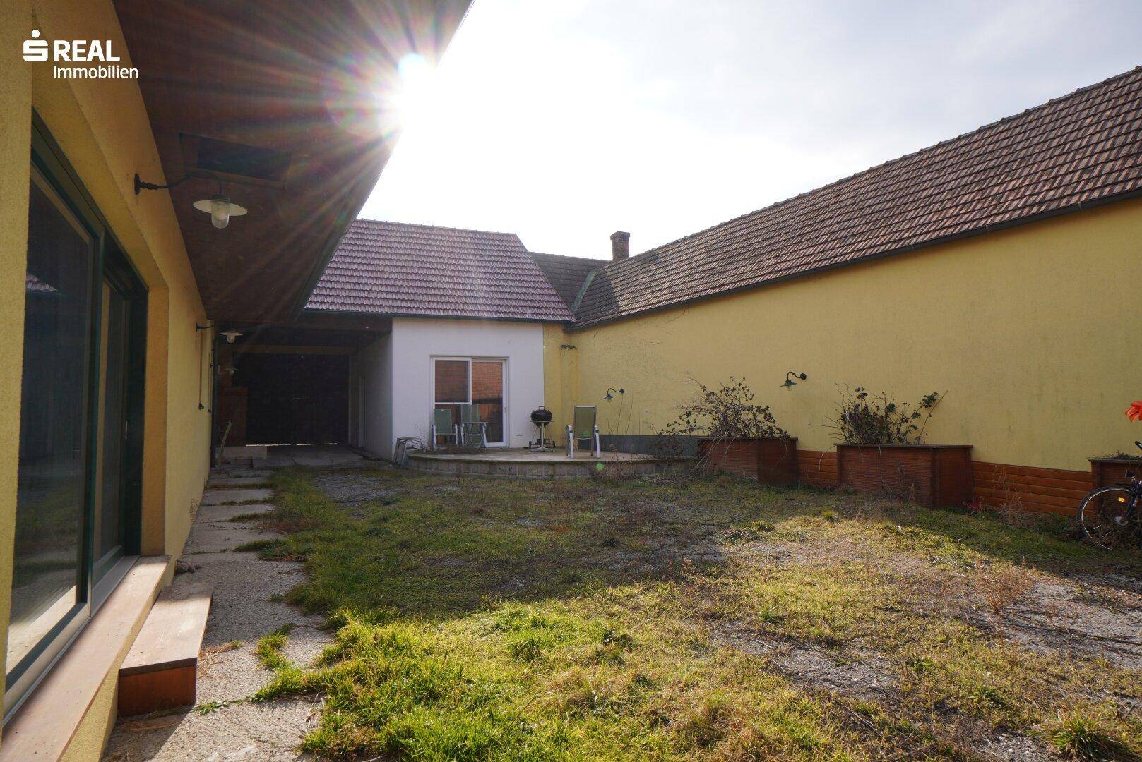 Sonniger Garten mit Rasenfläche und kleiner Terrasse, umgeben von Gebäuden.