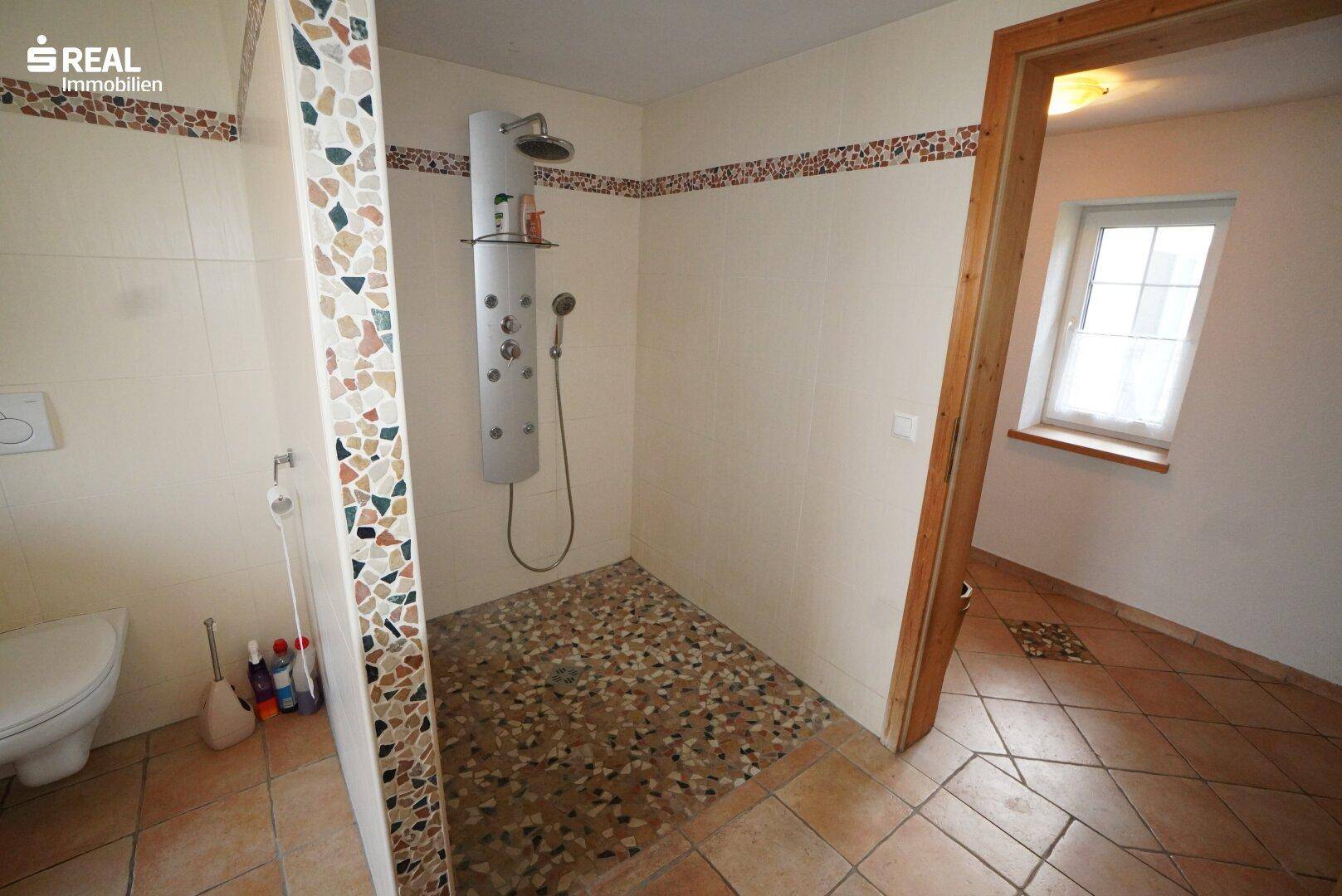Badezimmer mit ebenerdiger Dusche, WC und Fenster für Tageslicht.