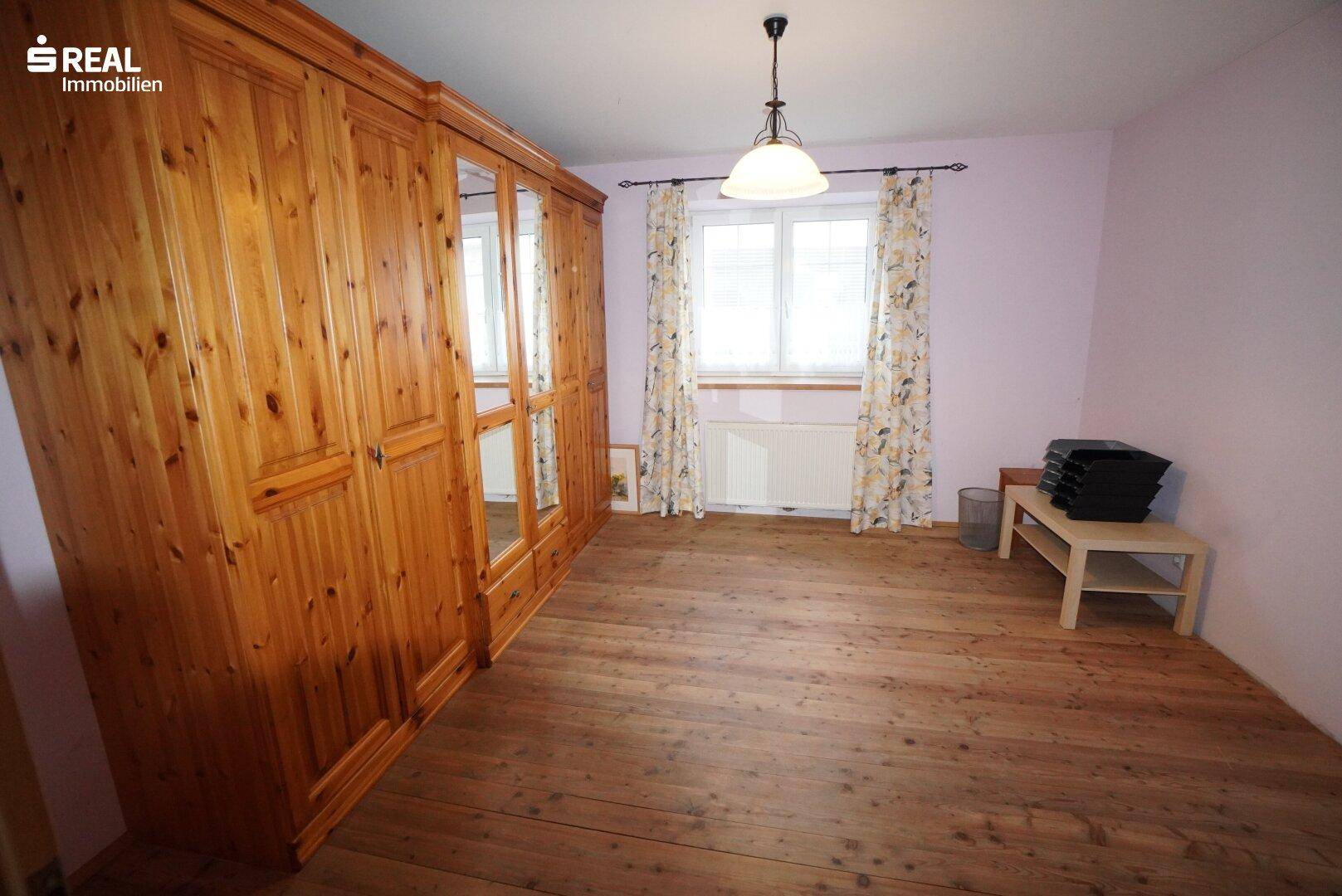 Schlafzimmer mit Holzboden, großem Holzschrank und Fenster mit Vorhängen.