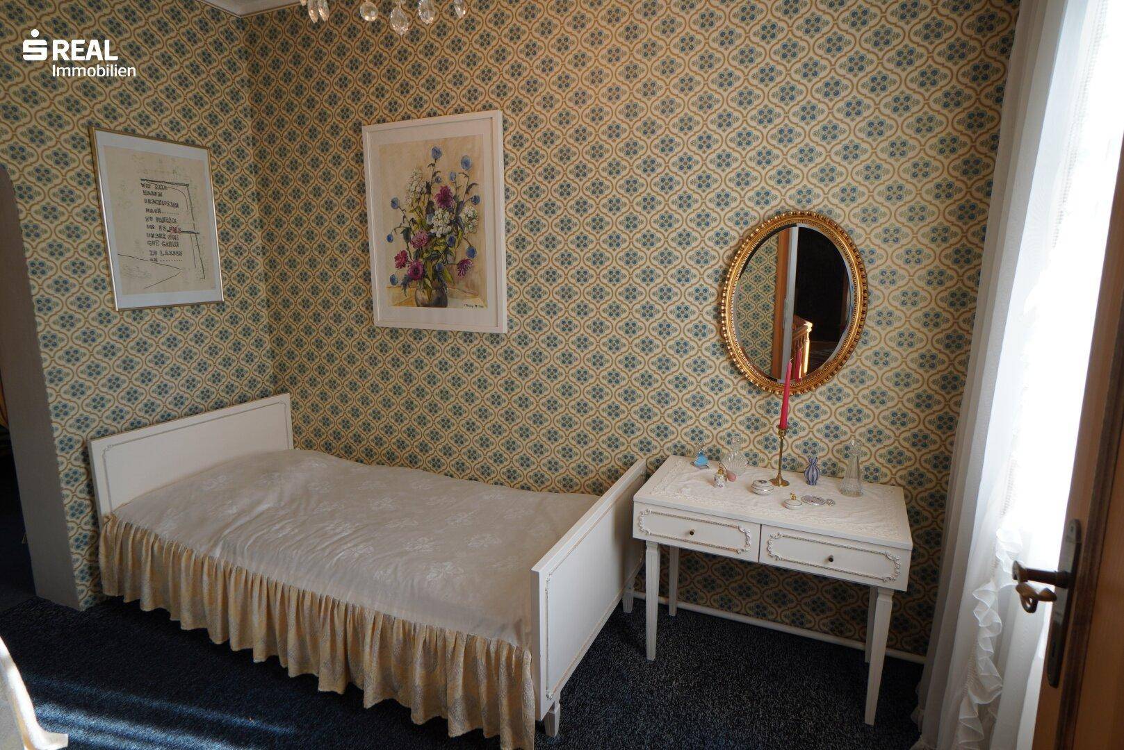 Schlafzimmer mit gemusterter Tapete, Einzelbett und einem kleinen Schminktisch.