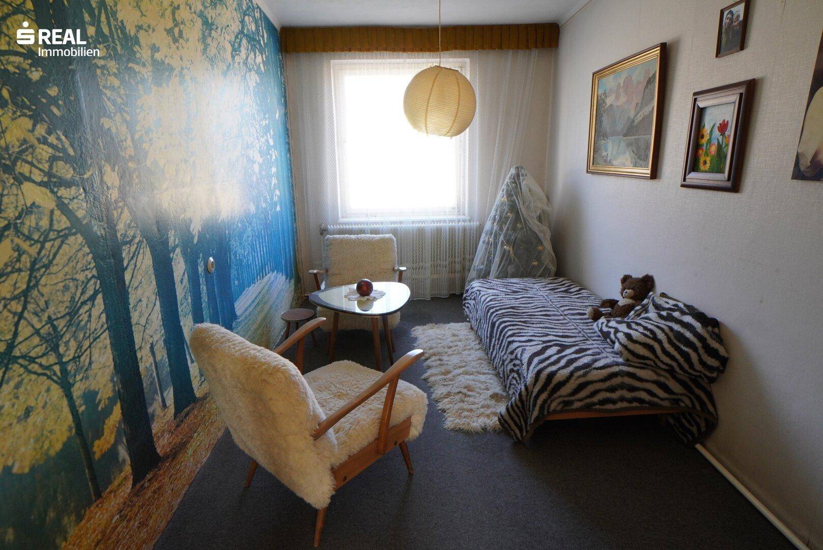 Schlafzimmer mit auffälliger Wandtapete, Einzelbett und zwei bequemen Sesseln am Fenster.