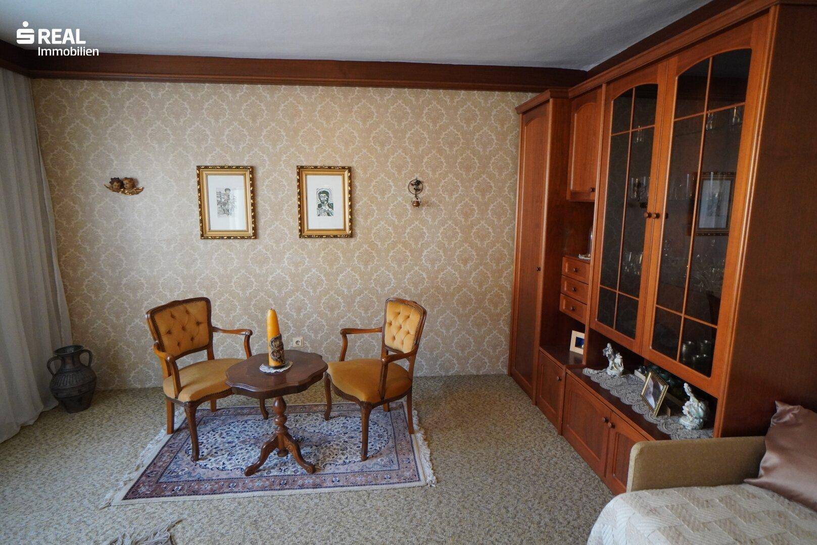 Wohnzimmer mit gemusterter Tapete, einem großen Holzschrank und Teppichboden.