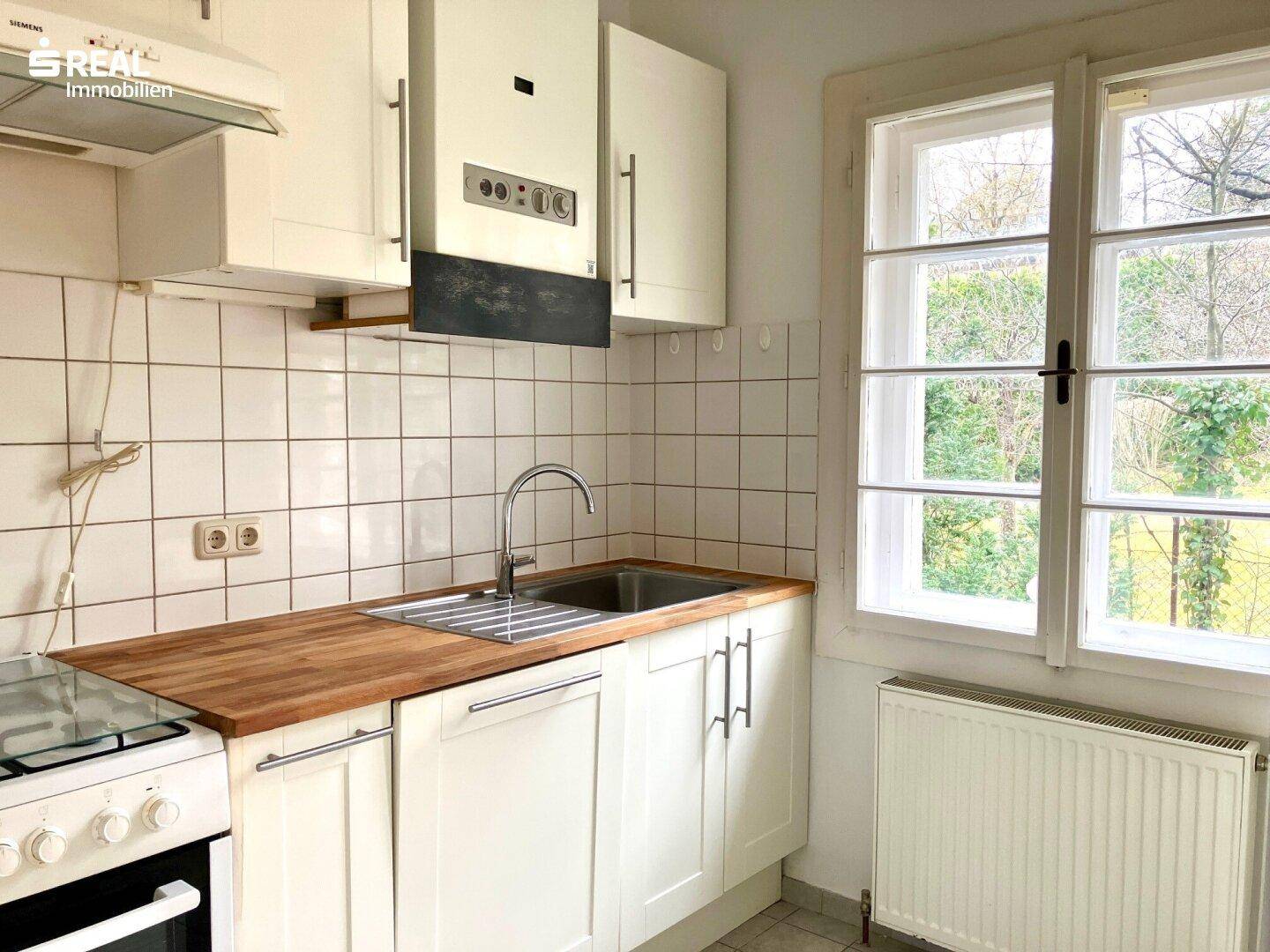 Kleine Küche mit weißen Schränken, Holz-Arbeitsplatte, Spüle und Fenster mit Blick ins Grüne.