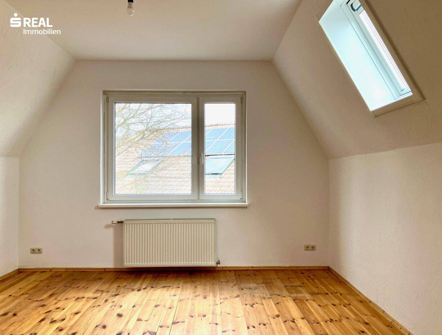 Helles Dachzimmer mit Holzboden, weißen Wänden und einem großen Fenster sowie einem Dachfenster.