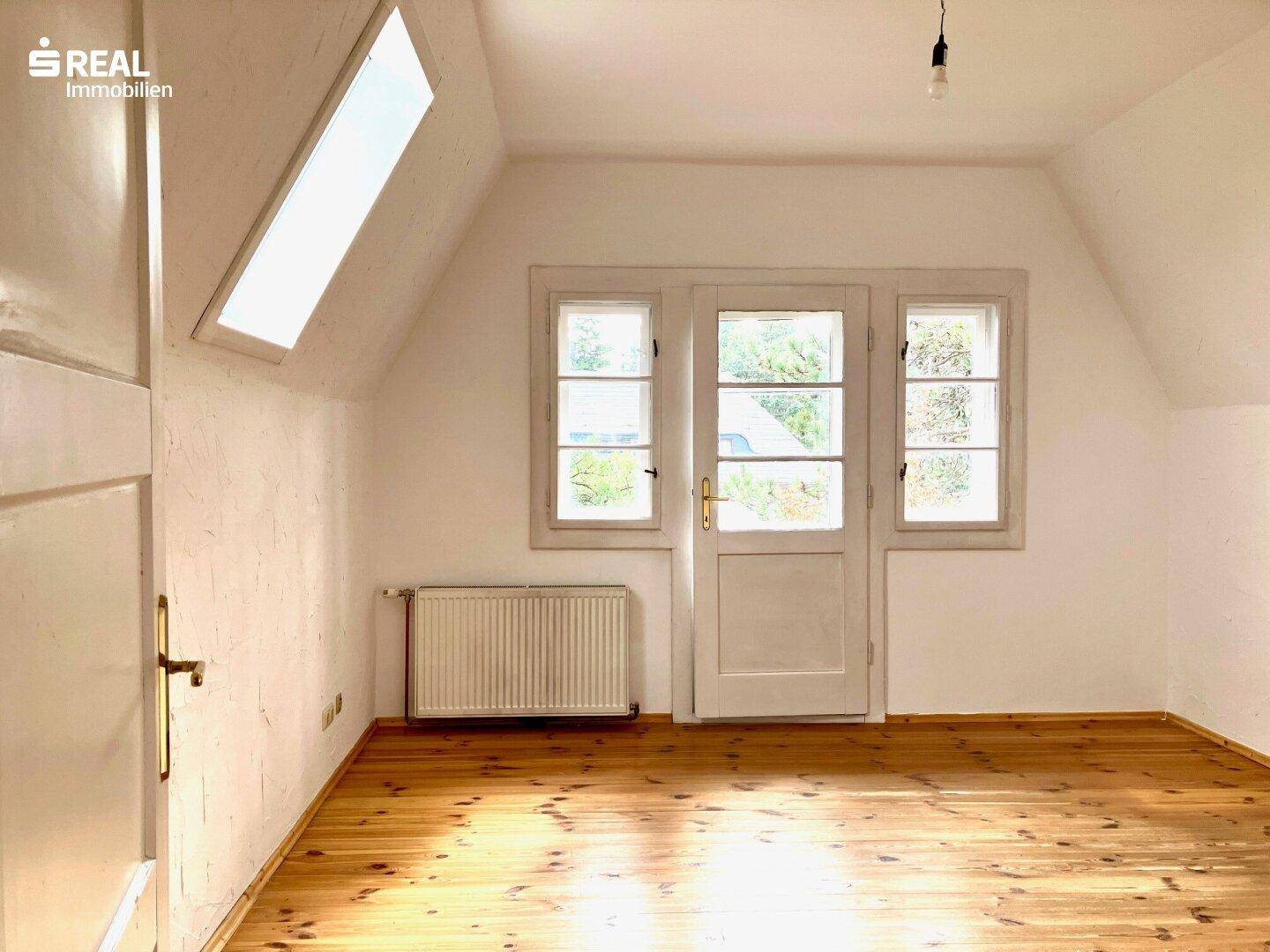 Dachzimmer mit Holzboden, weißen Wänden, Dachfenster und Zugang zu einem Balkon oder einer Terrasse.