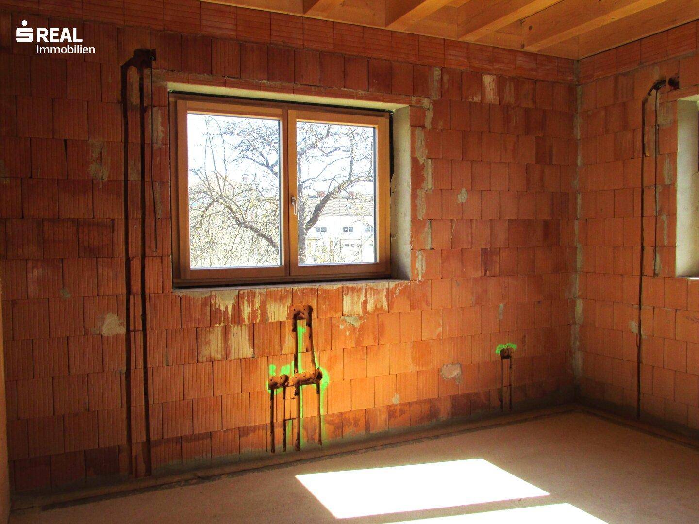 Rohbauzimmer mit Fenster und vorbereiteten Leitungen in den Ziegelwänden, viel Tageslicht.