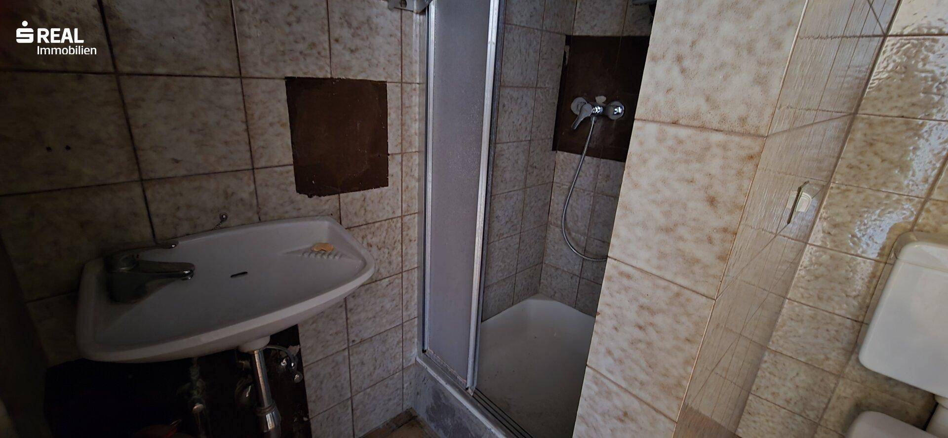 Badezimmer mit Fliesen, Waschbecken und einer Duschkabine, in einem älteren Zustand.