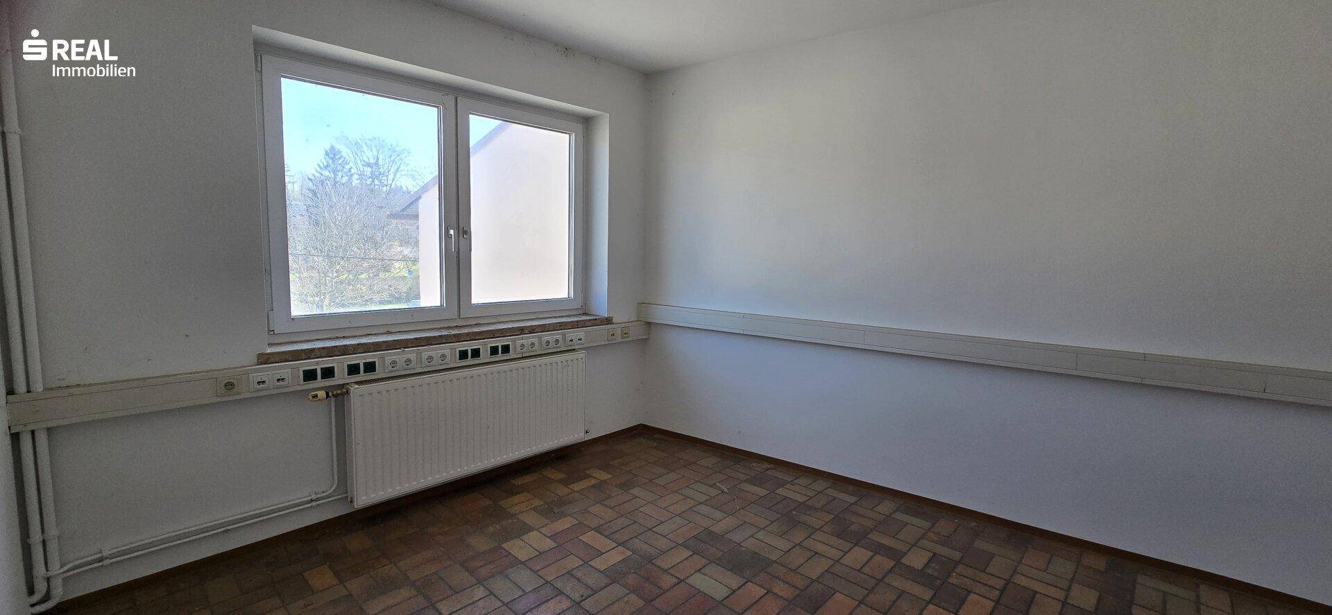 Helles Zimmer mit Parkettboden und einem großen Fenster, das einen Blick ins Grüne bietet.