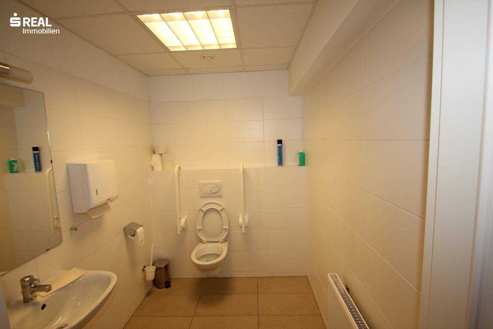 Barrierefreies WC mit Haltegriffen, wandhängender Toilette und Waschbecken.