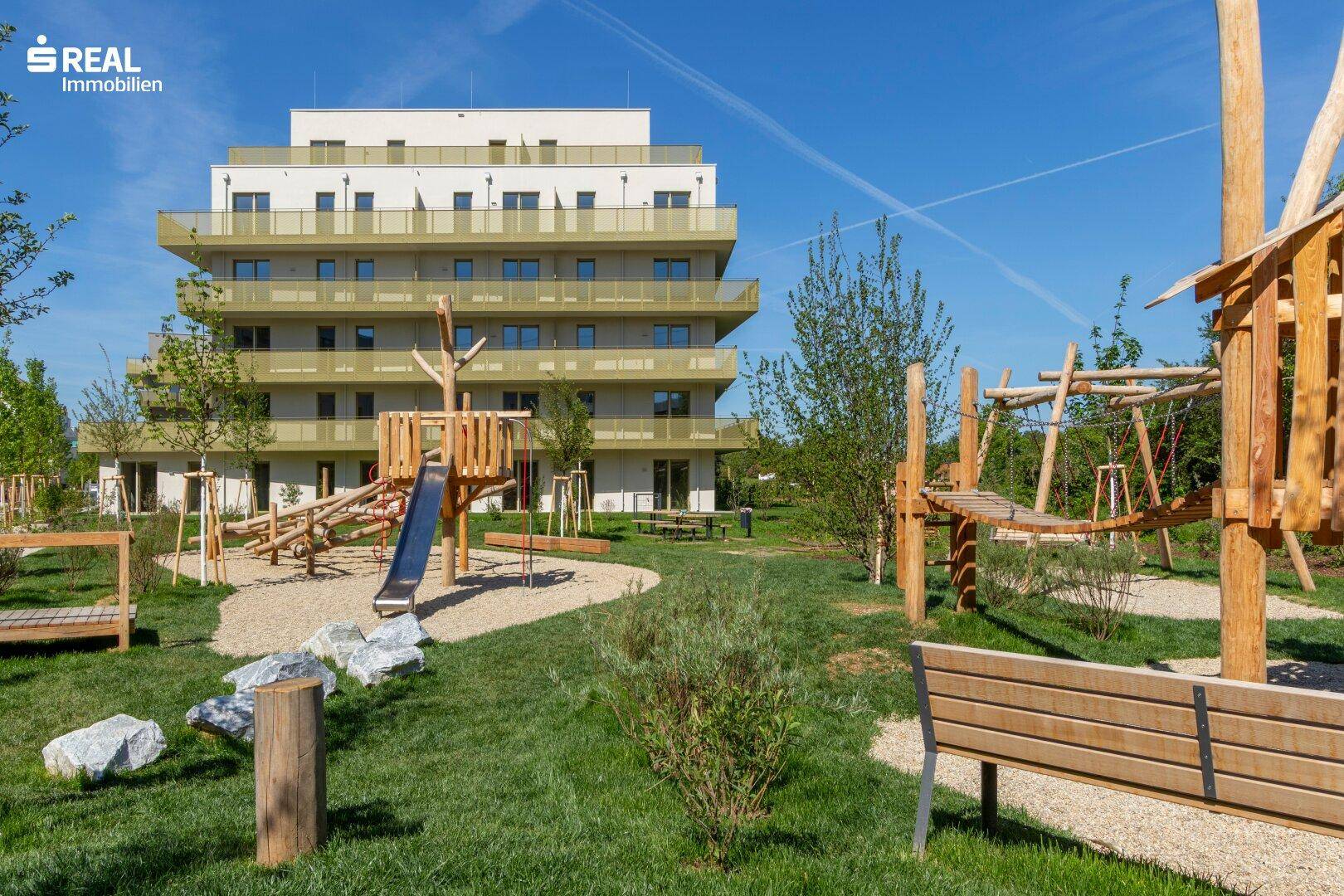 Modernes Wohngebäude mit Spielplatz im Vordergrund, umgeben von Grünflächen und Bäumen.