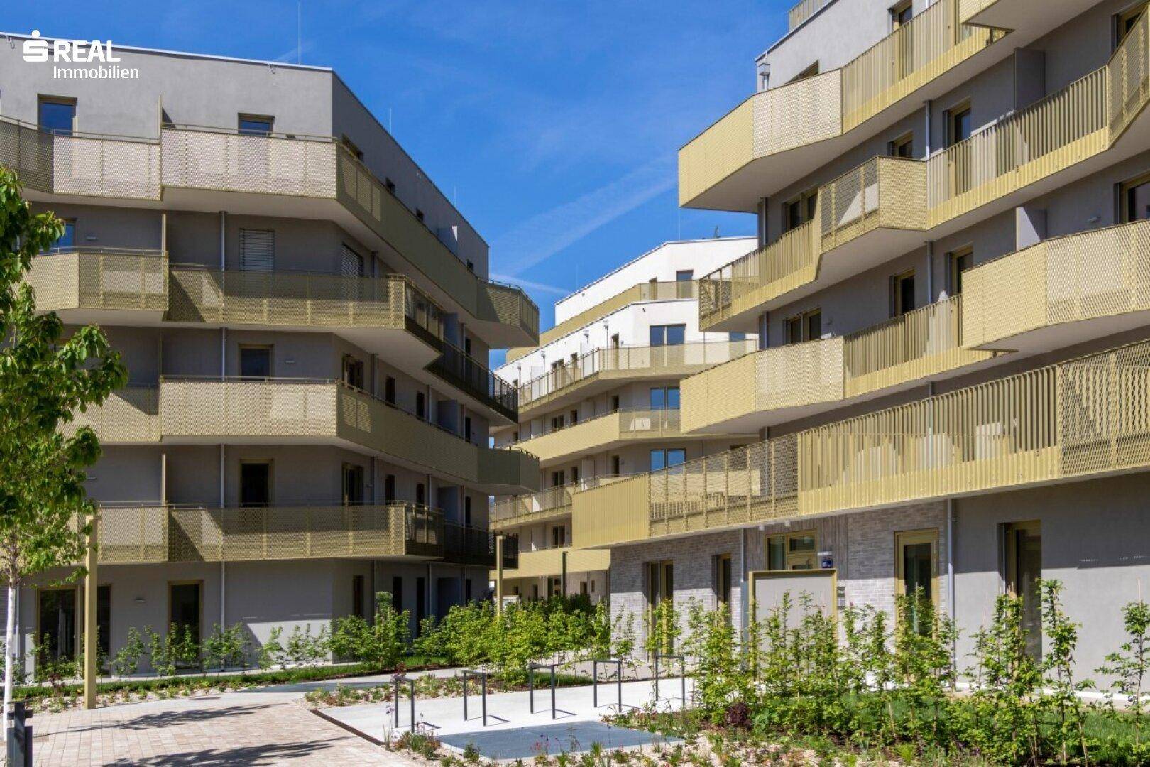 Zwei moderne Wohngebäude mit goldfarbenen Balkonen und großen Fenstern unter strahlend blauem Himmel.