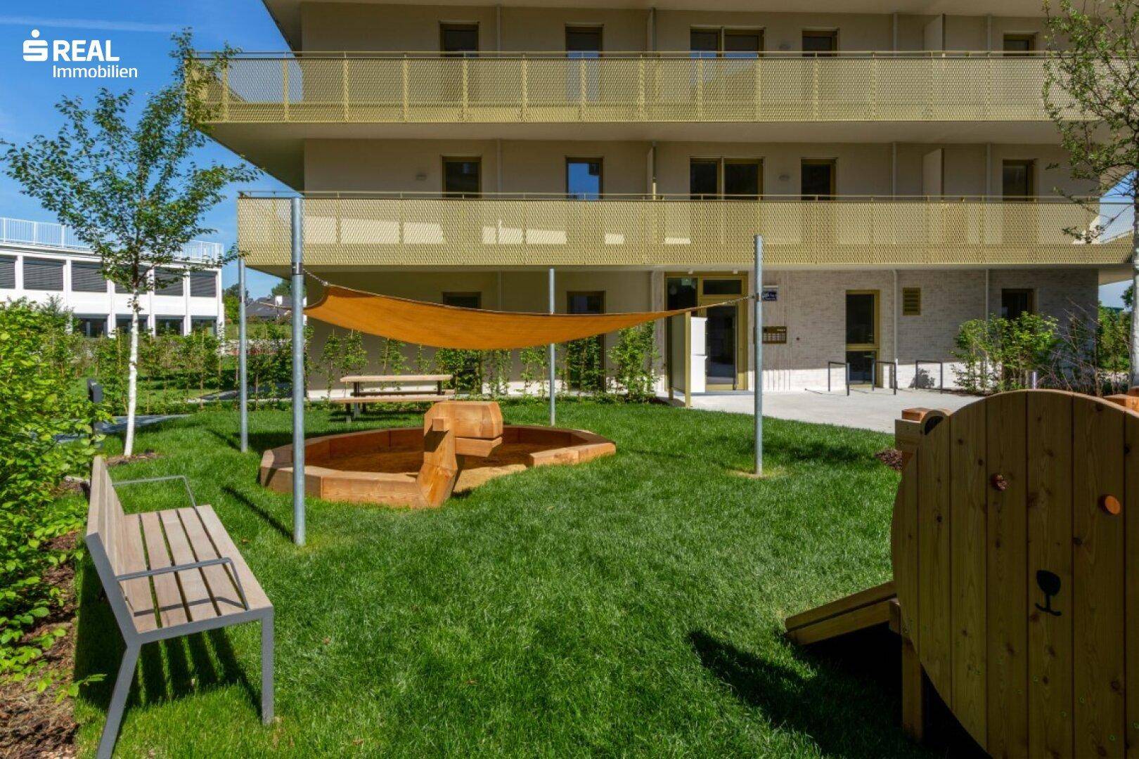 Grüner Gemeinschaftsgarten mit Spielbereich, Sandkasten und Sitzbänken für Kinder und Erwachsene.