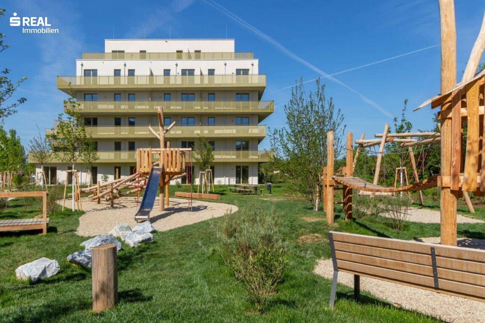 Moderne Wohnanlage mit großem, naturnahem Spielplatz im Vordergrund, ausgestattet mit Holzstrukturen und Rutschen.